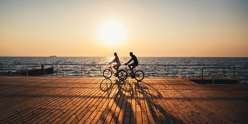 Couple of young hipsters cycling together at the beach at sunrise sky at wooden deck summer time.