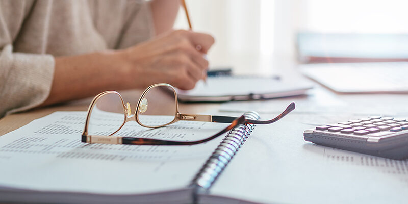 Eyeglasses and calculator close-up and accountant woman making marks in paper documents with modern laptop sitting at desk background next bright daylight window. Business, home finances money savings